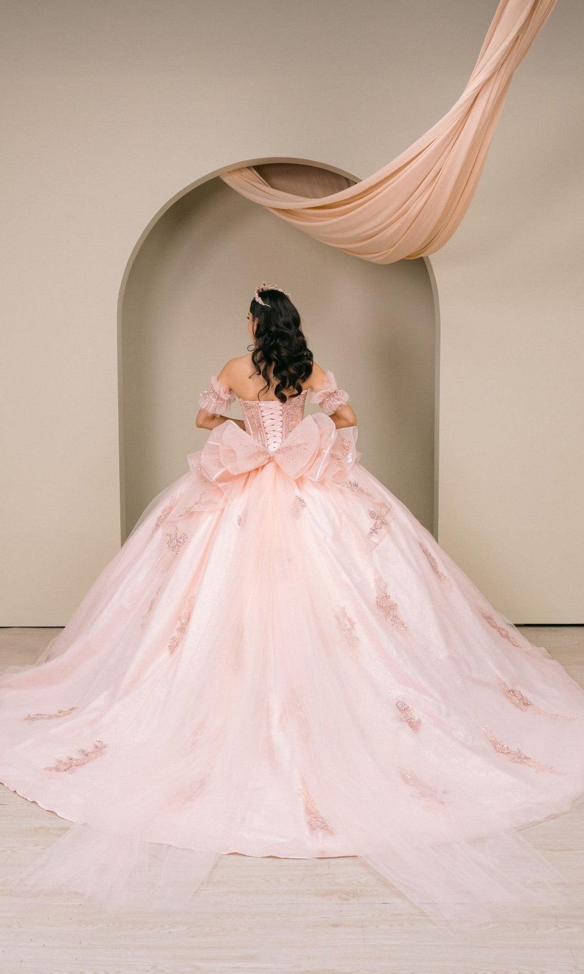 A woman wearing the Quinceanera Dress 8540JQ by Cinderella Couture, a blush pink gown with lace details, stands facing away in front of a beige arch adorned with draped fabric.